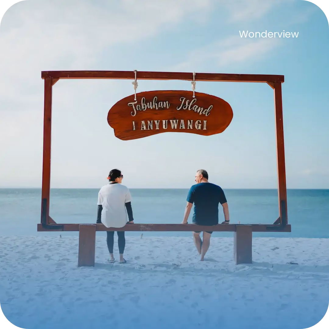 Dua wisatawan duduk di bangku Pantai Pulau Tabuhan dengan latar papan bertuliskan ‘Tabuhan Island Banyuwangi.