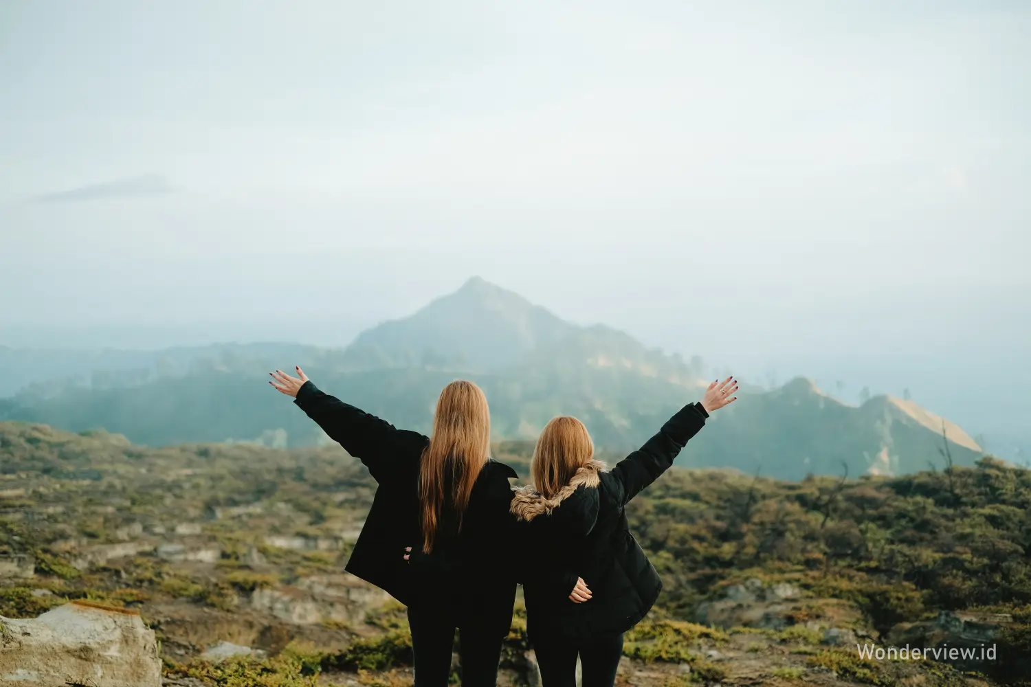 Dua perempuan berdiri membelakangi kamera sambil merentangkan tangan menghadap panorama pegunungan Kawah Ijen.