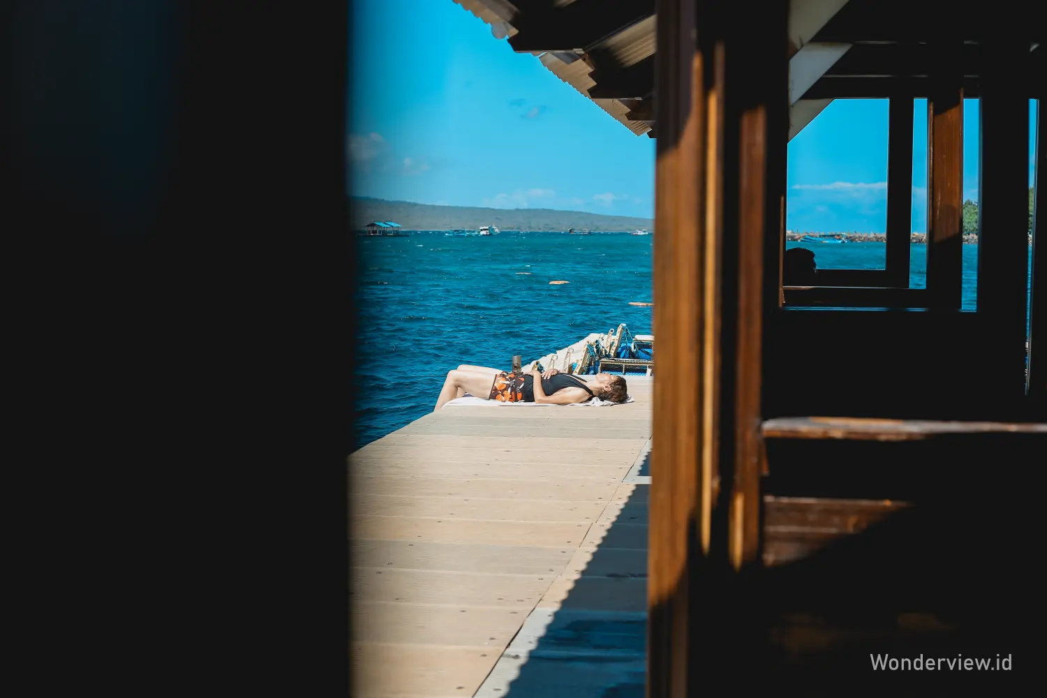 Wisatawan bersantai di dermaga Bangsring Banyuwangi dengan latar laut biru dan perahu di kejauhan.
