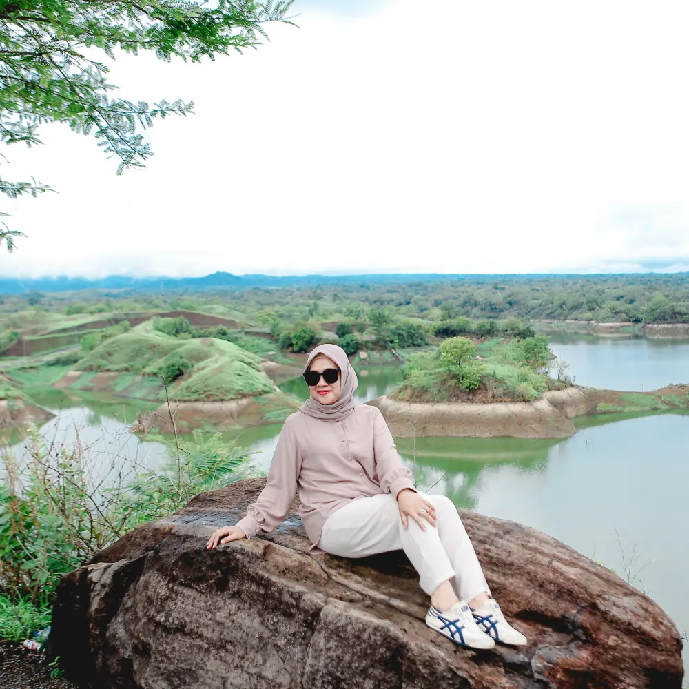 Wisatawan berpose di atas batu besar dengan pemandangan Waduk Bajul Mati yang dikelilingi perbukitan hijau di Banyuwangi.