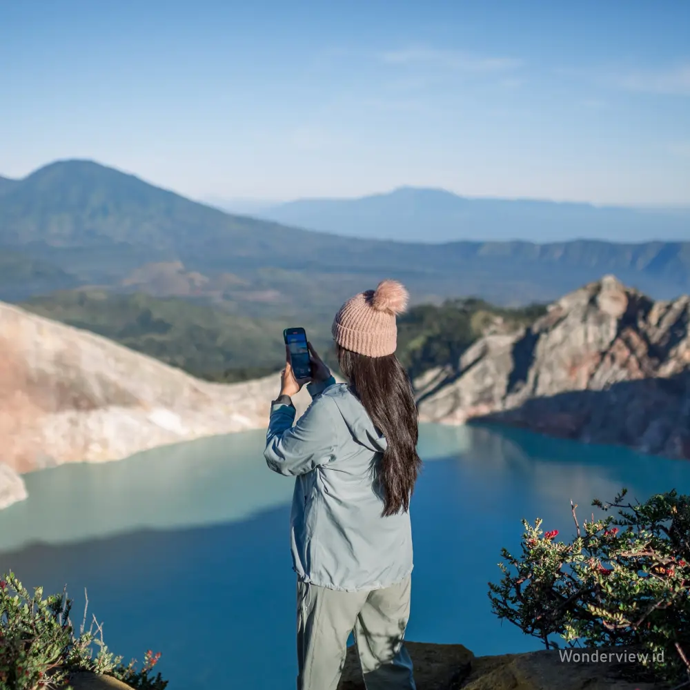 Seorang wisatawan memotret pemandangan Danau Kawah Ijen yang berwarna biru toska dari tepi tebing.