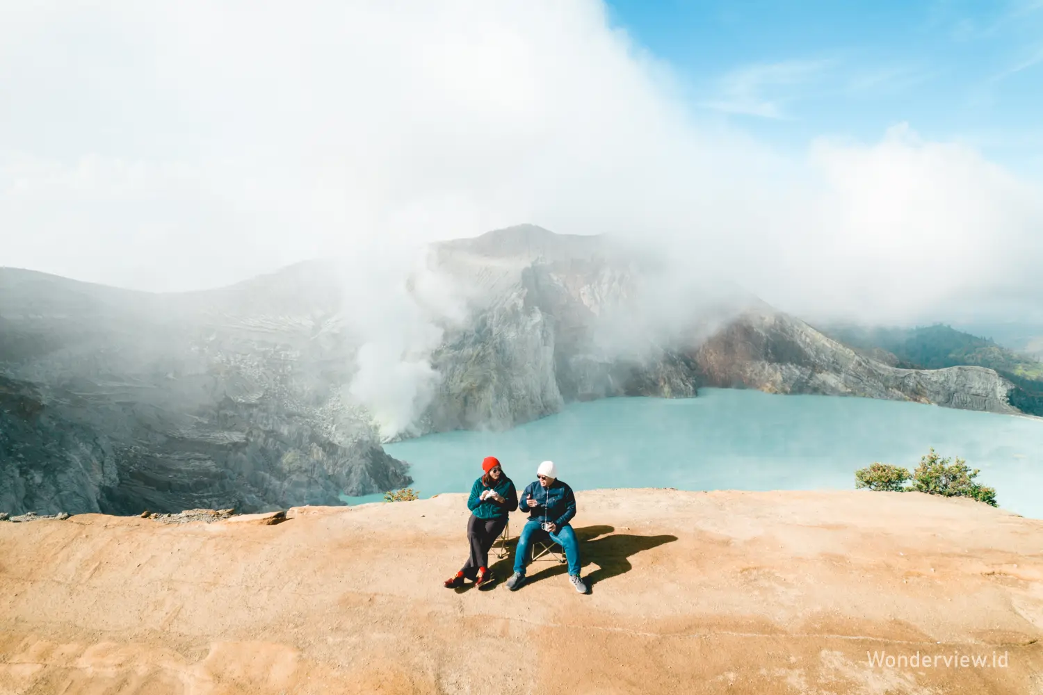Dua wisatawan duduk di tepi kawah Ijen sambil menikmati pemandangan danau biru dan asap belerang.