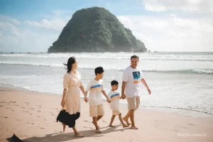 Keluarga berjalan menyusuri tepi Pantai Pulau Merah dengan latar bukit ikonik dan ombak tenang.