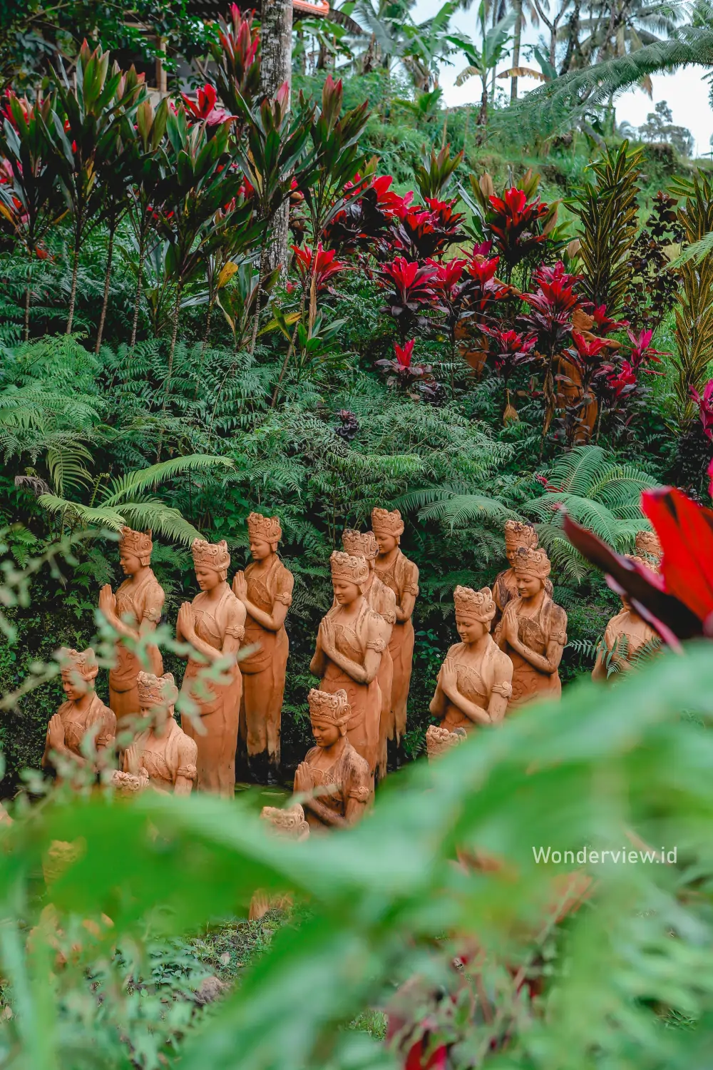 Deretan patung gandrung terakota berdiri di tengah taman hijau dengan tanaman tropis yang rimbun.