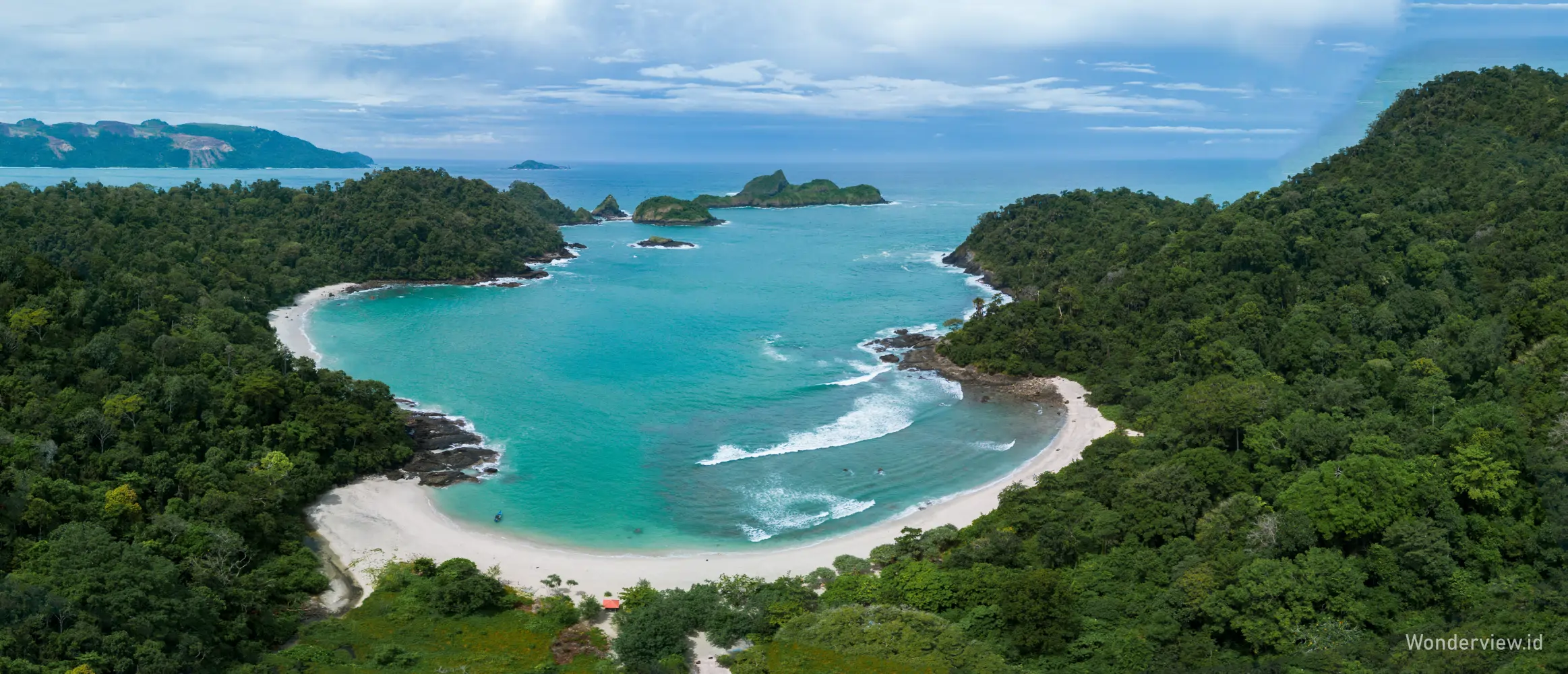 Pemandangan udara Pantai Wedi Ireng dengan laut biru jernih, teluk berpasir putih, dan hutan hijau yang mengelilinginya.