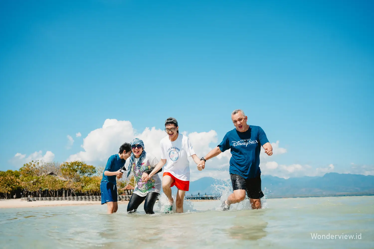 Empat orang berjalan ceria di perairan dangkal Pulau Menjangan dengan latar langit biru dan pepohonan pesisir.