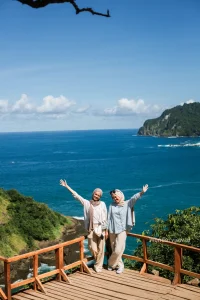 Dua wisatawan berpose di deck kayu dengan latar laut biru dan tebing hijau di South Sea Banyuwangi.