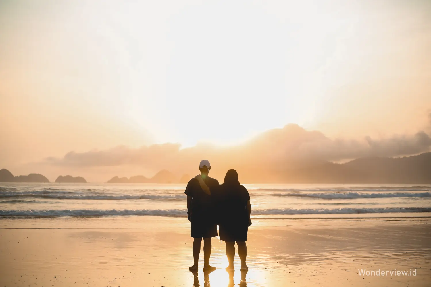 Pasangan menikmati matahari terbenam di Pantai Pulau Merah Banyuwangi dengan siluet ombak dan langit keemasan.