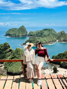 Dua wisatawan berpose di viewpoint Green Island Banyuwangi dengan latar tebing hijau dan laut biru.