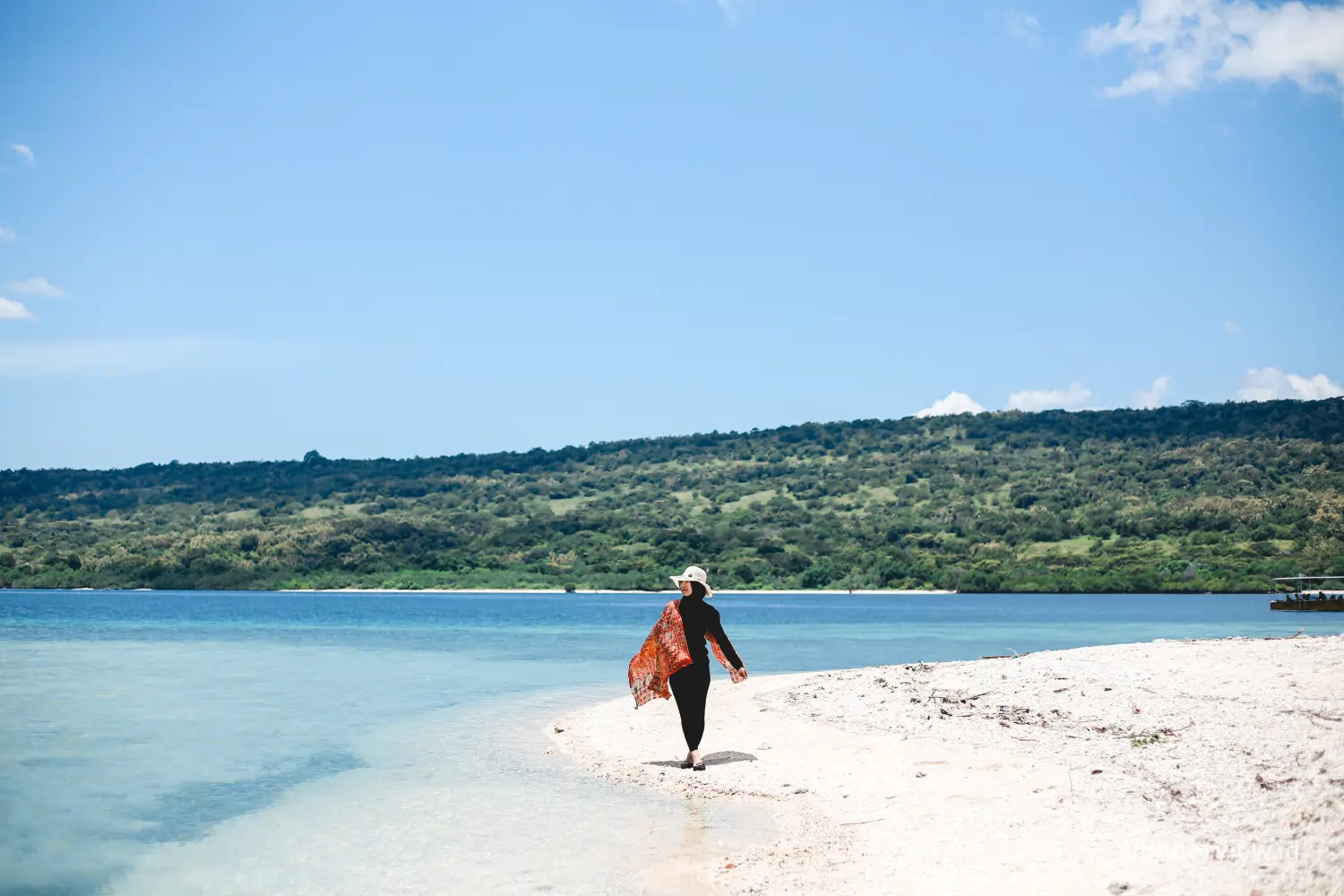 Wisatawan berjalan di tepi pantai Pulau Menjangan dengan air laut jernih dan bukit hijau di kejauhan.