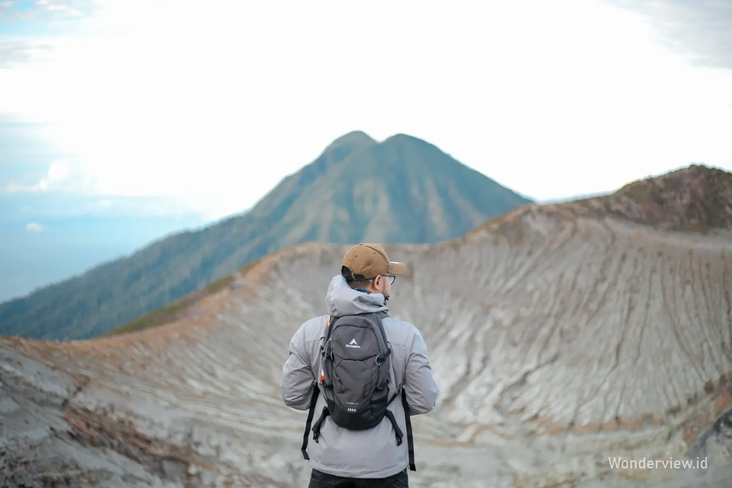 Pendaki berdiri di tepi kawah Ijen Banyuwangi dengan latar pegunungan dan dinding kawah bertekstur.