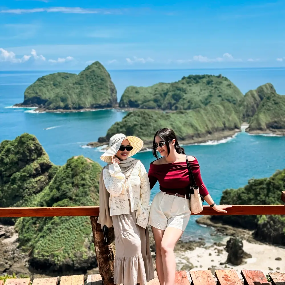 Dua wisatawan berpose di Puncak Green Island Banyuwangi dengan latar tebing hijau dan laut biru.