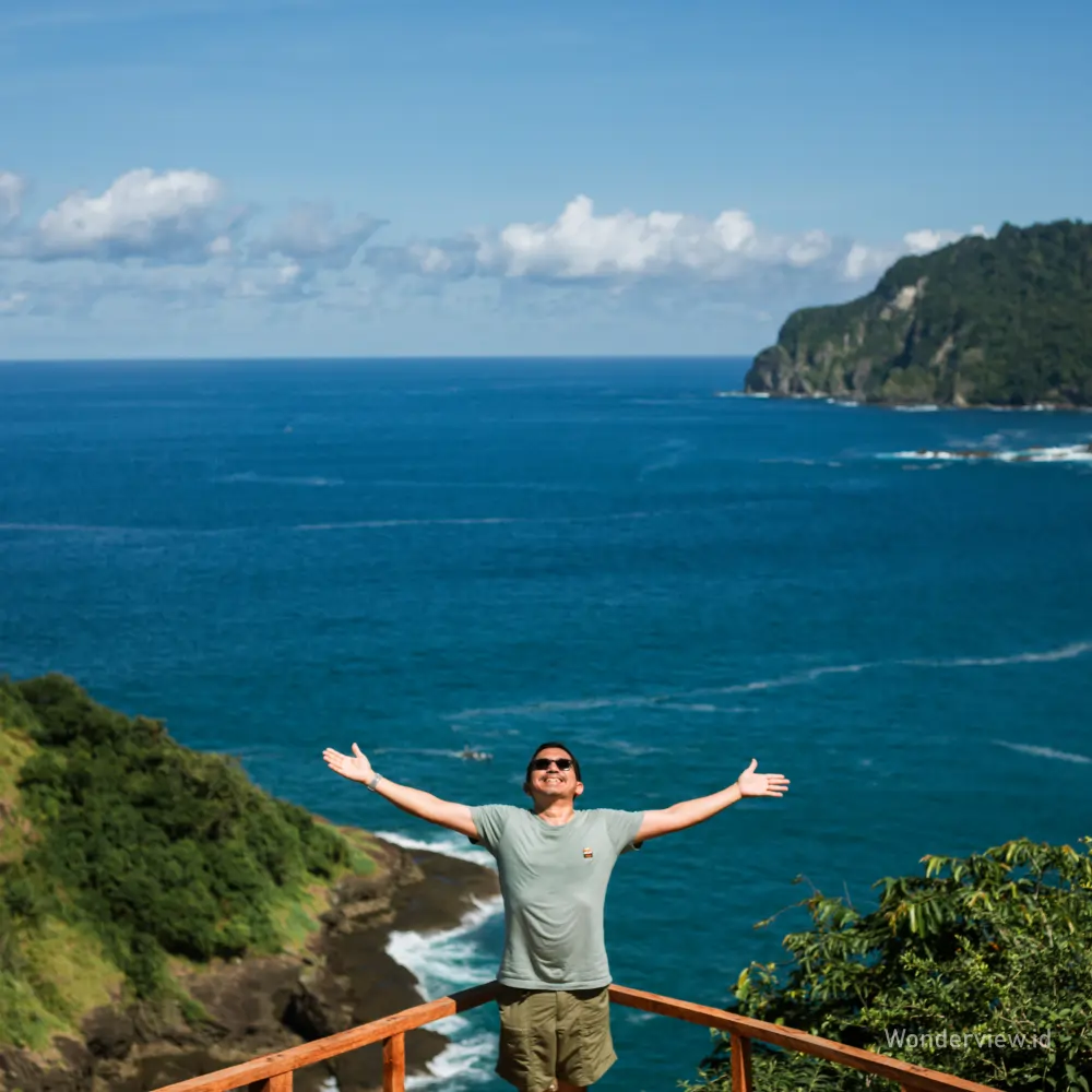 Seorang wisatawan berdiri di atas viewpoint Green Island Banyuwangi dengan latar laut biru dan tebing hijau.